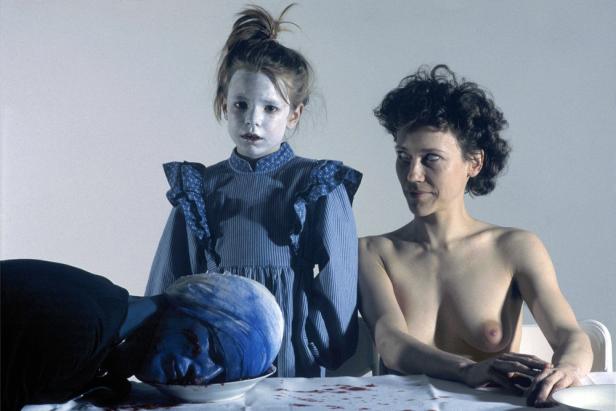 Eine blass geschminkte junge Frau, eine barbusige Frau und ein blauer Kopf auf einem Teller an einem Tisch.