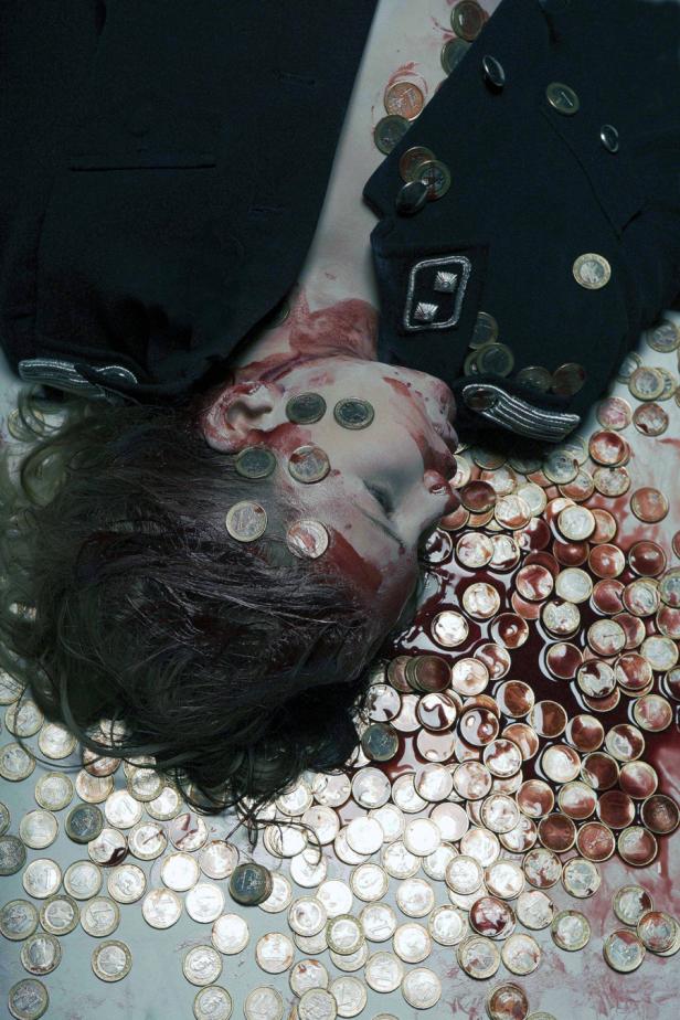 Eine tote Person in Uniform liegt auf dem Boden, bedeckt mit Blut und Euromünzen.