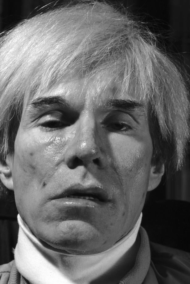 Ein Schwarzweiß-Porträt von Andy Warhol mit weißblondem Haar und einem ernsten Gesichtsausdruck.