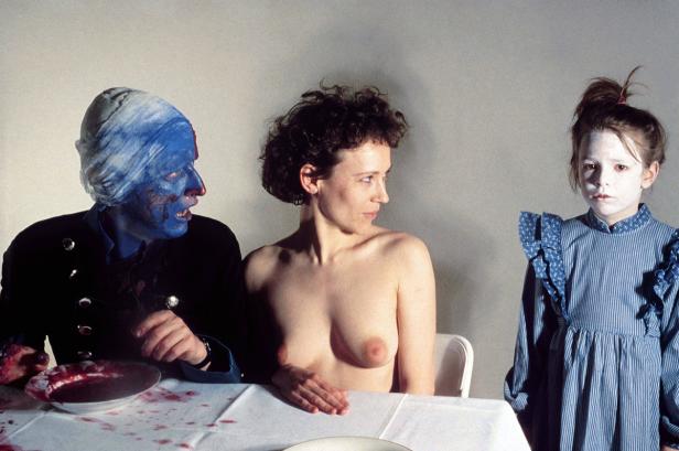 Ein Mann mit blau bemalten Gesicht, eine Frau mit nacktem Oberkörper und ein Mädchen sitzen an einem Tisch.