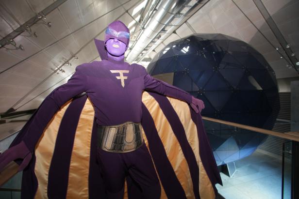 Eine Person in einem lila Superheldenkostüm mit einem goldenen Umhang steht in einem Gebäude.
