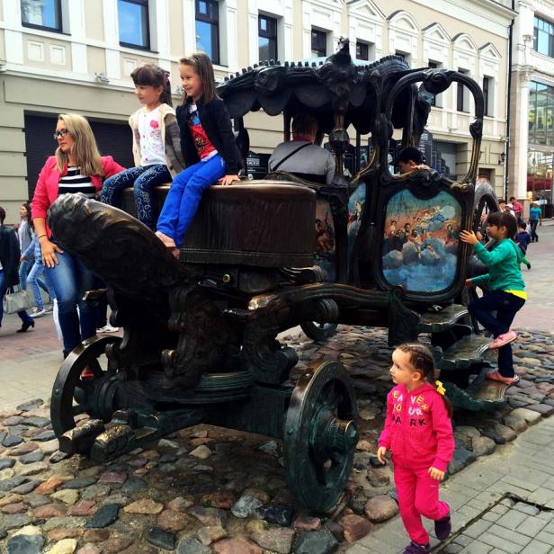 Mehrere Kinder spielen an einer Kutschenskulptur auf einem gepflasterten Platz.