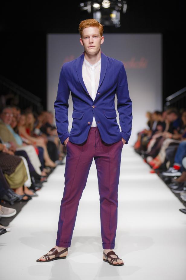Ein rothaariger Mann präsentiert ein blaues Jackett und weinrote Hosen auf einem Laufsteg.
