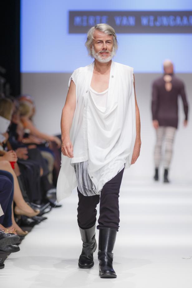 Ein Mann mit grauem Haar und Bart läuft auf einem Laufsteg in einem weißen Outfit.