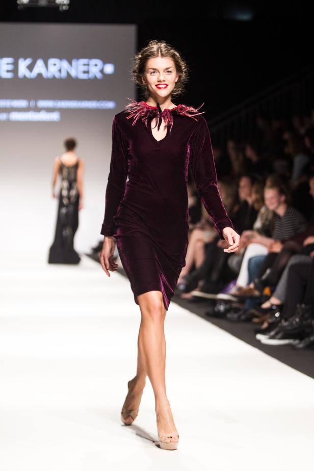 Eine Frau präsentiert ein burgunderrotes Samtkleid mit Federn auf einem Laufsteg.