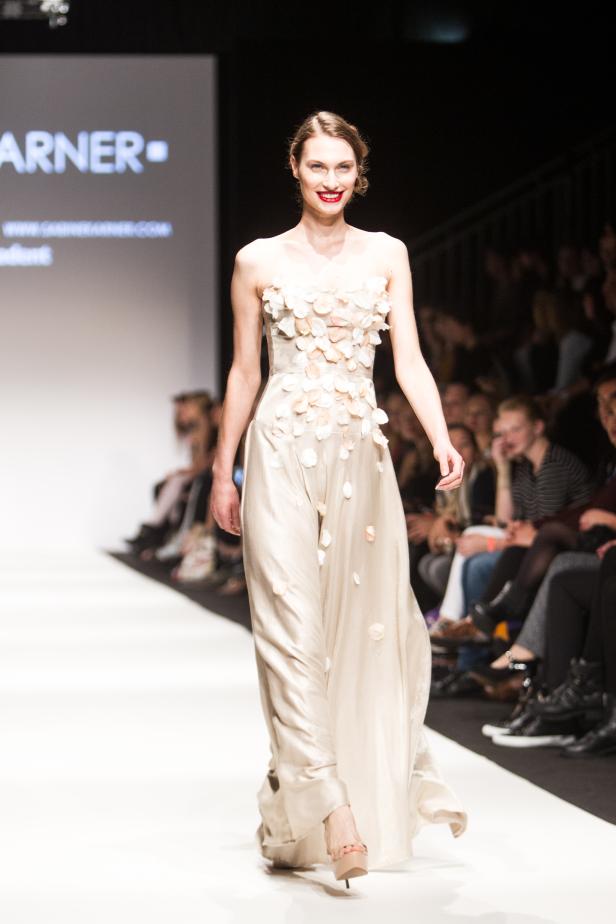 Eine Frau präsentiert ein langes, helles Kleid mit Blütenapplikationen auf einem Laufsteg.