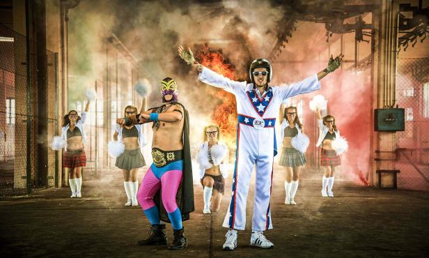 Ein Wrestler mit Maske, ein Mann im Superheldenkostüm und Cheerleader posieren vor Rauchschwaden.