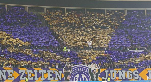 Zuschauer im Stadion halten goldene und lila Folien hoch, um ein Muster zu erzeugen.