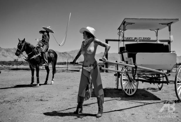 Eine Frau im Cowboy-Outfit posiert vor einem Wagen, während ein Mann auf einem Pferd im Hintergrund steht.