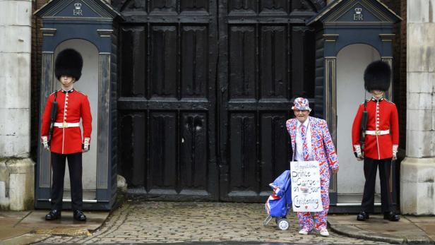 Zwei königliche Gardisten stehen neben einem Mann im Union-Jack-Anzug vor einem Tor.