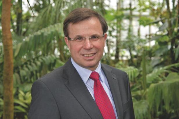 Ein Mann mit Brille und Anzug steht vor tropischen Pflanzen.