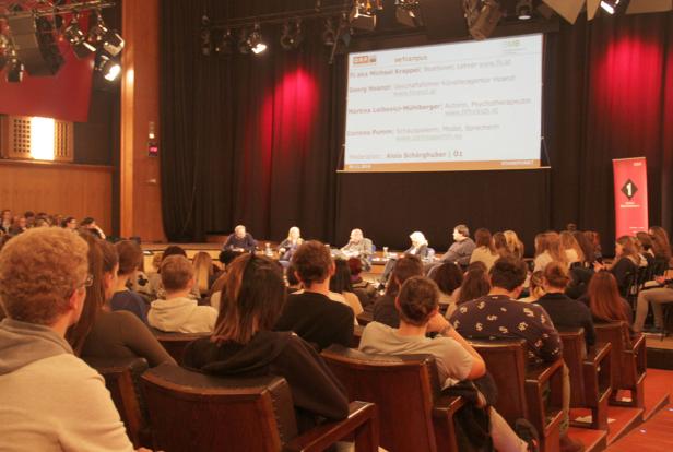Publikum sitzt bei einer Podiumsdiskussion mit mehreren Teilnehmern auf der Bühne.