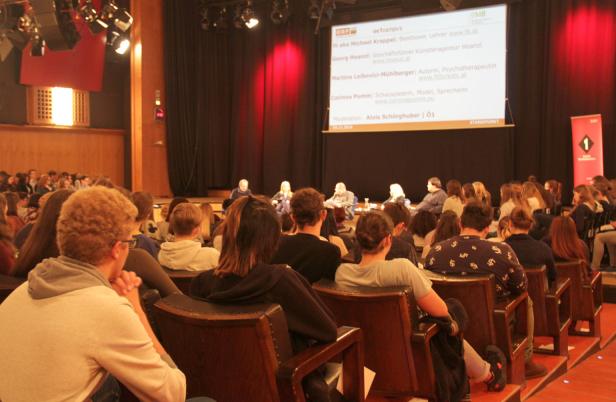 Zuschauer sitzen in einem Saal und schauen einer Podiumsdiskussion zu.