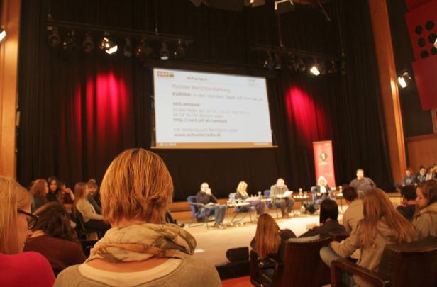 Publikum sitzt vor einer Bühne mit einer Leinwand und einer Podiumsdiskussion.