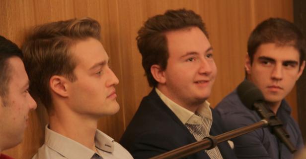 Vier junge Männer sitzen nebeneinander vor einer Holzwand, ein Mikrofon ist im Vordergrund zu sehen.