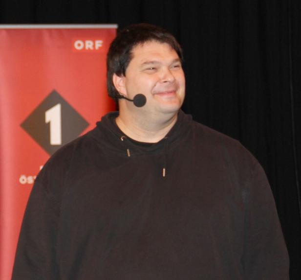 Ein Mann mit Mikrofon lächelt vor einem ORF-Banner.