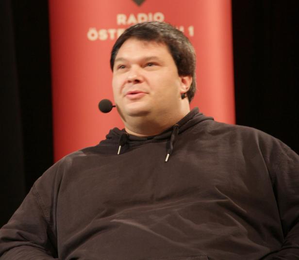Ein Mann mit Mikrofon vor einem roten Hintergrund mit der Aufschrift „Radio Österreich 1“.