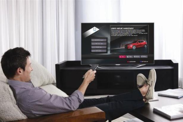 Ein Mann entspannt auf einem Sofa und sieht sich den neuen Mazda 3 im Fernsehen an.