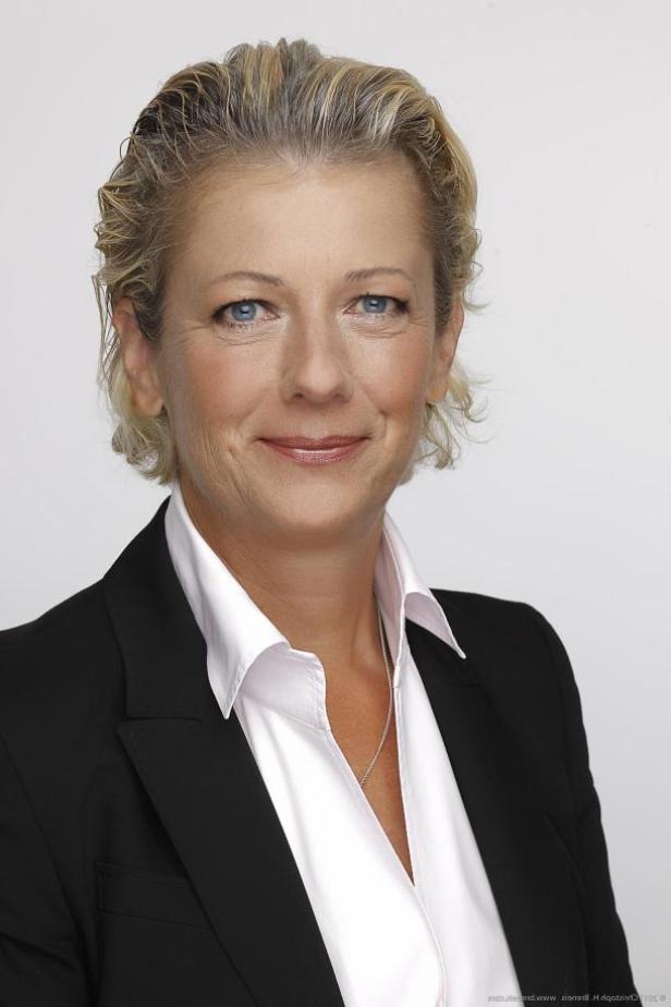 Porträt einer Frau mit blonden Haaren, die eine schwarze Jacke und eine weiße Bluse trägt.
