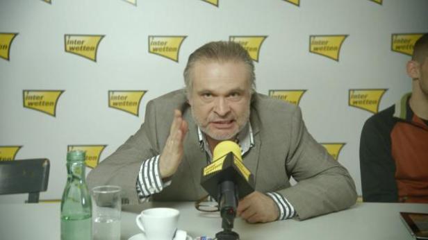 Ein Mann gestikuliert vor einem Mikrofon mit dem „Interwetten“-Logo im Hintergrund.