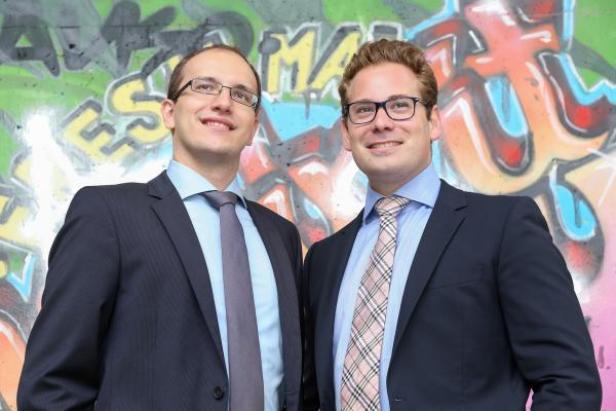 Zwei Männer in Anzügen stehen vor einer Wand mit Graffiti.