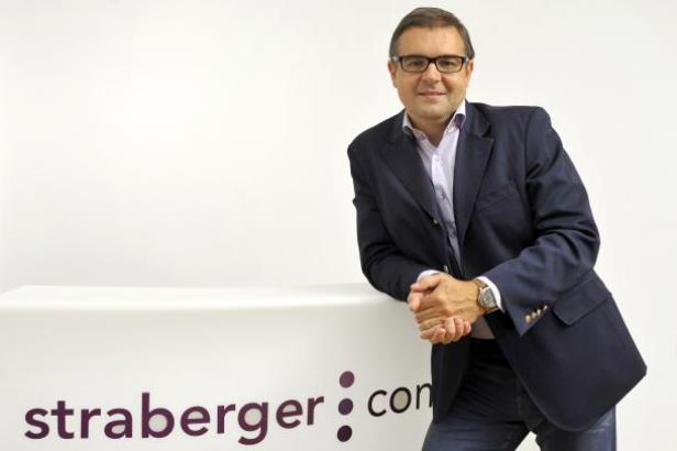 Ein Mann mit Brille und Sakko lehnt an einem Tisch mit dem Straberger-Logo.