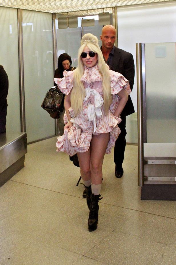 Lady Gaga in einem rosa Kleid mit Rüschen und Sonnenbrille, begleitet von einem Bodyguard.