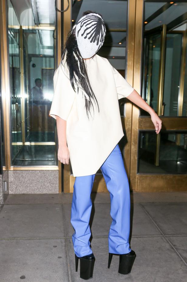 Eine Person mit einer weißen Maske, einem beigen Oberteil und blauen Hosen vor einem Gebäude.
