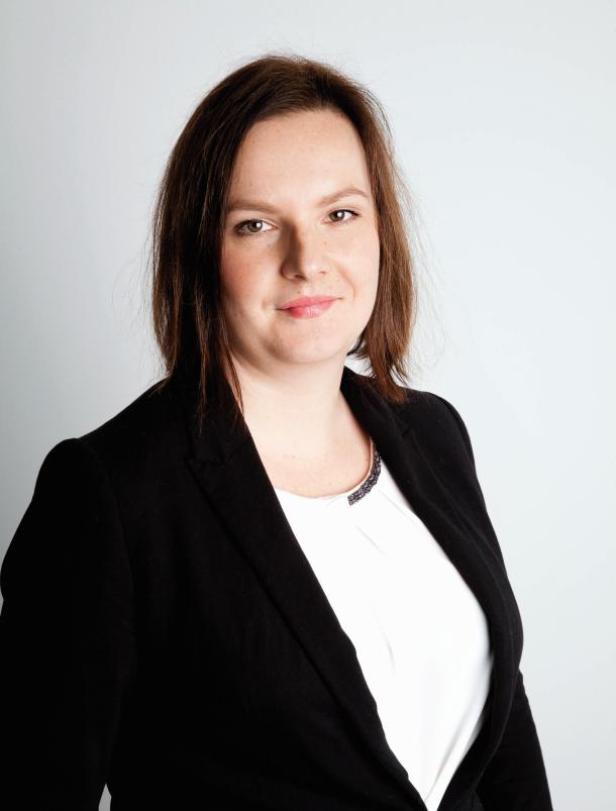 Eine Frau mit braunen Haaren und einem schwarzen Blazer lächelt in die Kamera.