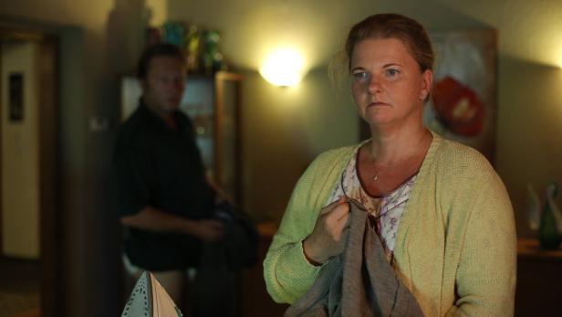 Eine Frau mit Strickjacke hält ein Kleidungsstück in der Hand, während ein Mann im Hintergrund steht.