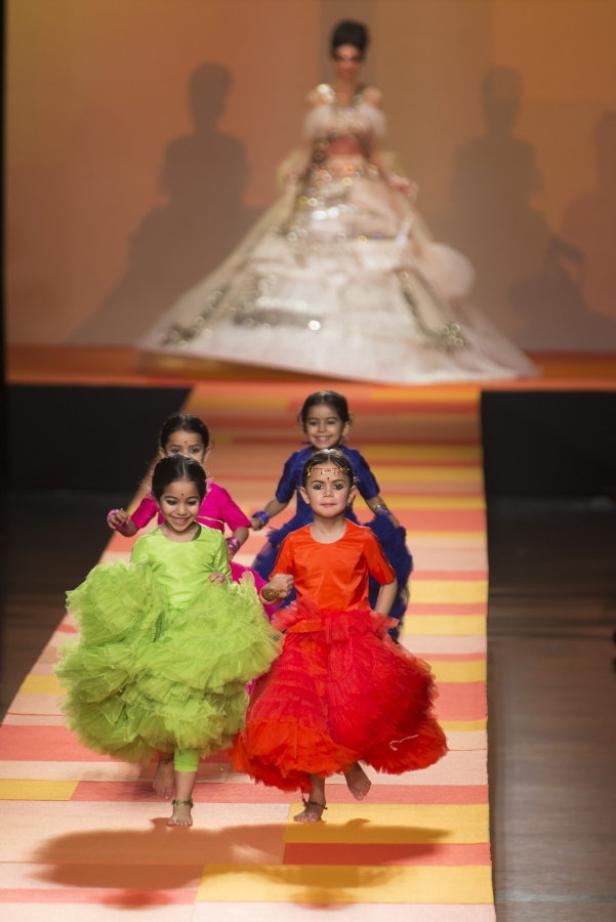 Eine Gruppe junger Mädchen in farbenfrohen Kleidern läuft barfuß über einen Laufsteg.