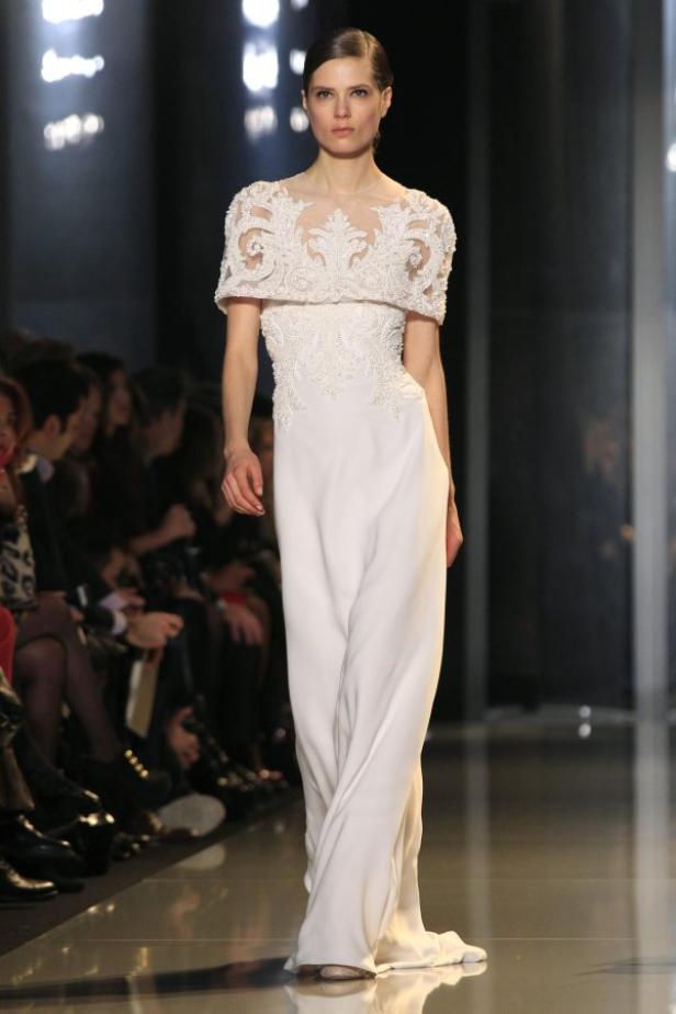 Eine Frau präsentiert ein langes, weißes Kleid mit Spitze auf einem Laufsteg.