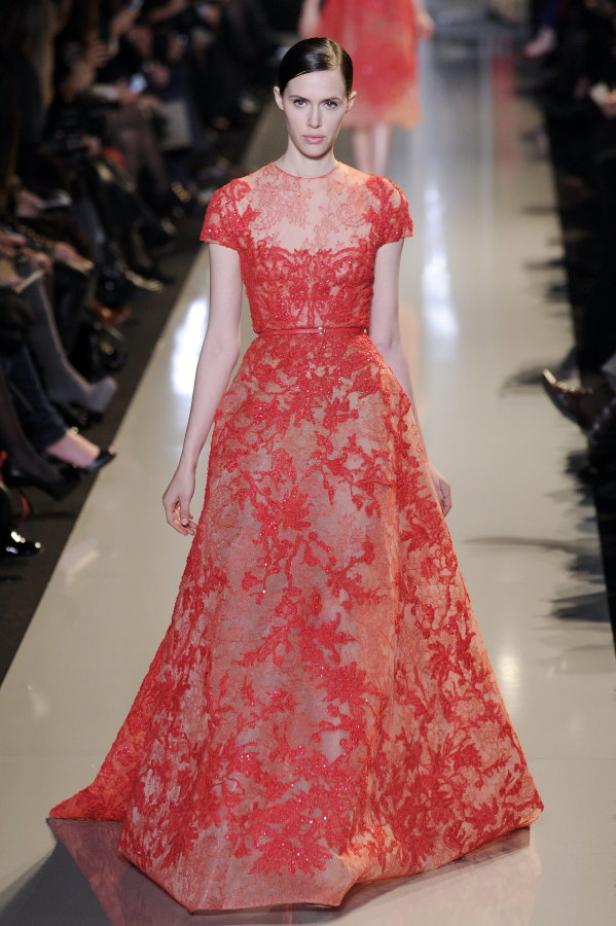 Ein Model präsentiert ein langes, rotes Kleid mit floraler Spitze auf einem Laufsteg.