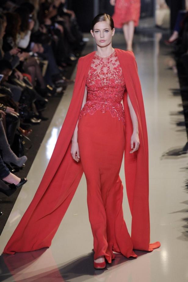 Eine Frau in einem roten, bodenlangen Kleid mit Cape auf einem Laufsteg.