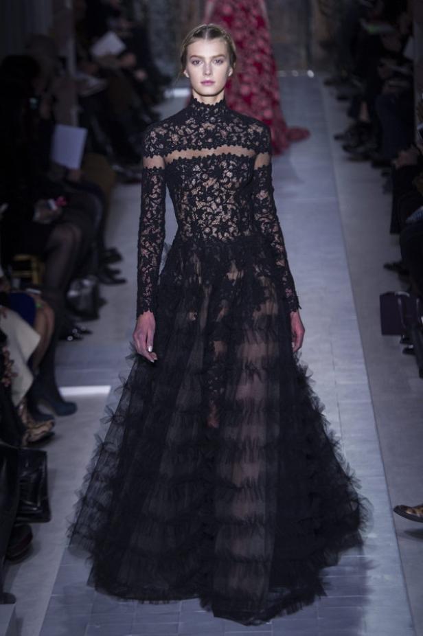 Ein Model präsentiert ein langes, schwarzes Spitzenkleid mit Rüschen auf einem Laufsteg.