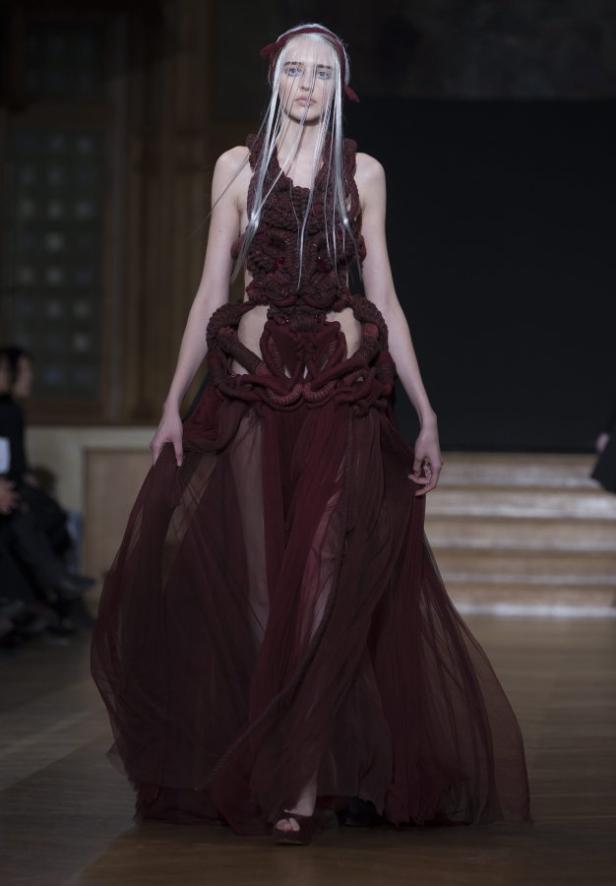 Ein Model präsentiert ein extravagantes, bordeauxrotes Kleid auf dem Laufsteg.