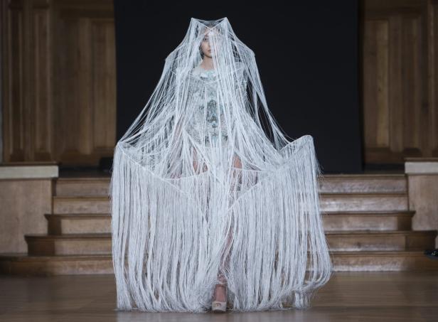 Ein Model präsentiert ein extravagantes Kleid mit langen, weißen Fransen auf einem Laufsteg.