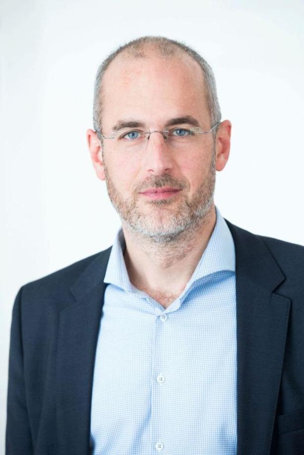 Ein Mann mit Brille, Hemd und Sakko vor einem weißen Hintergrund.