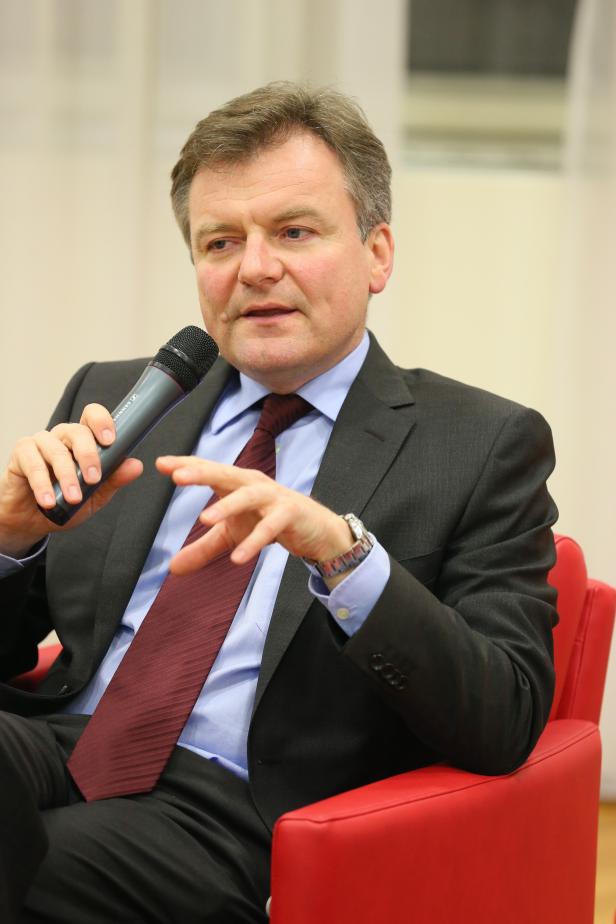Ein Mann im Anzug spricht in ein Mikrofon, während er auf einem roten Sessel sitzt.