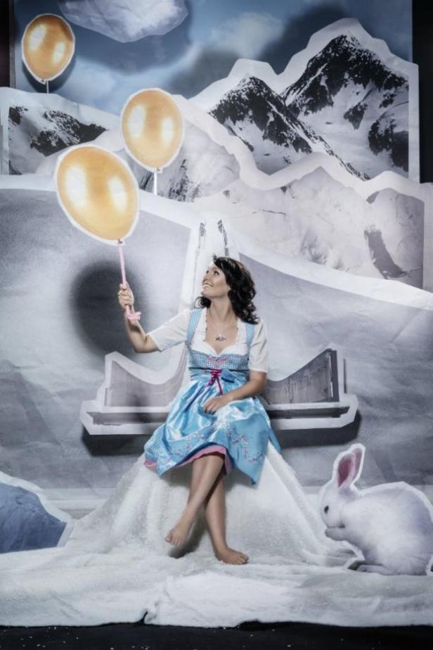 Jacqueline Seifriedsberger - Skispringerin. Auf einem Alice-in-Winterland-Motiv. Indiz dafür: der weisse Hase. (c: zooom.at - markus berger) Eine Frau im Dirndl sitzt vor einer Bergkulisse und hält Luftballons in der Hand. Ein Hase sitzt daneben.