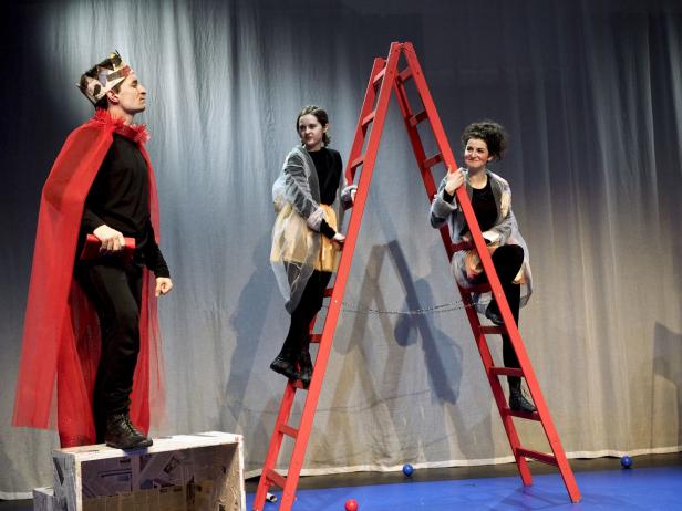 Drei Schauspieler auf einer Bühne mit einer roten Leiter und einem König.