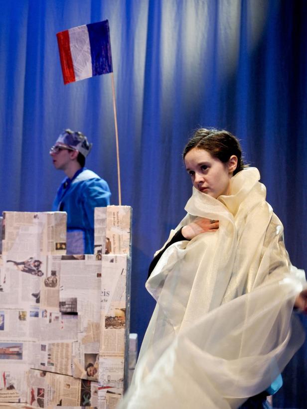 Eine Frau in einem weißen Gewand steht vor einer Burgkulisse mit einer französischen Flagge im Hintergrund.