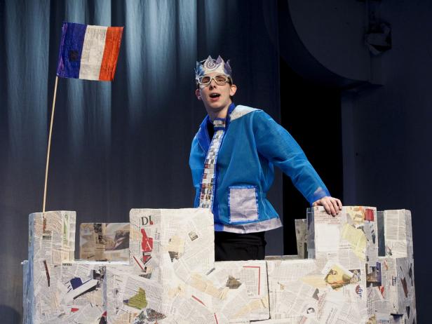 Ein junger Mann in einem blauen Kostüm und einer Krone steht hinter einer Burg aus Zeitungspapier mit einer französischen Flagge.