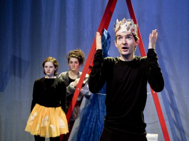 Ein Mann mit einer Krone aus Zeitungspapier gestikuliert auf einer Theaterbühne.