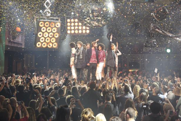 Eine Gruppe von Models steht auf einem Laufsteg unter Konfettiregen vor einem Publikum.