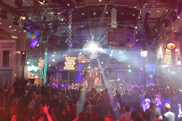 Eine große Menschenmenge feiert unter einer Discokugel und bunten Lichtern.