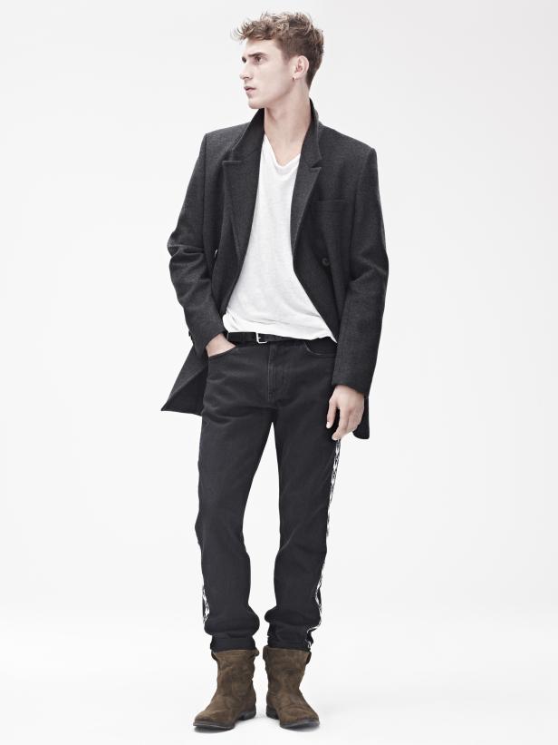 Ein Mann mit dunkler Jeans, Stiefeln und einem grauen Mantel posiert vor einem weißen Hintergrund.