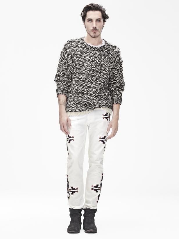 Ein Mann mit gemustertem Pullover und verzierten weißen Hosen steht vor einem weißen Hintergrund.