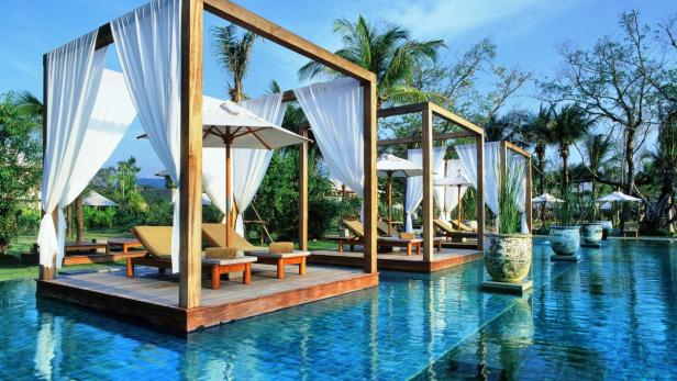 Mehrere Cabanas mit Liegestühlen stehen an einem Pool in einem tropischen Resort.