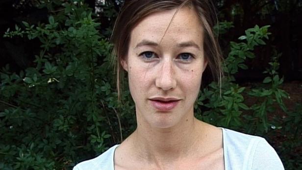 Eine Frau mit hellbraunen Haaren und blauen Augen vor einem grünen Busch.